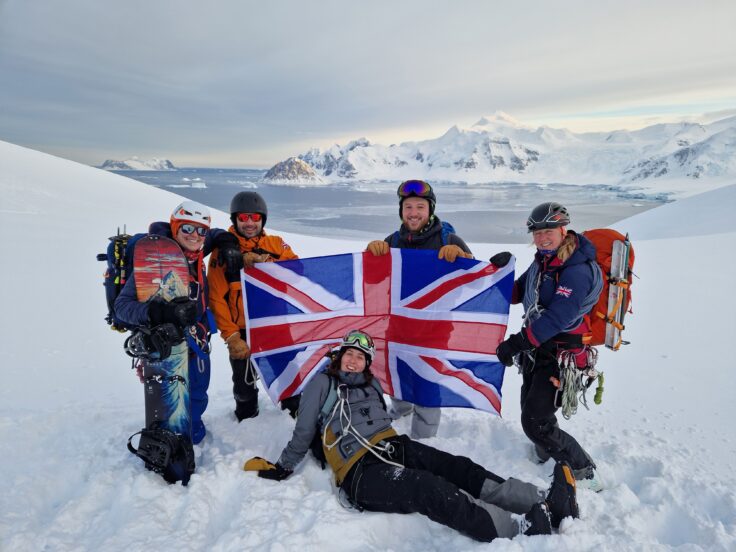 A group of five people dressed in ski clothes stand in the snow holding a Union Jack flag. There is water and a snowy mountain behind them.